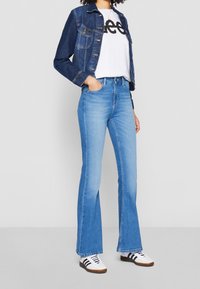 Blue denim flared jeans paired with a white graphic tee and a dark denim jacket. Footwear includes white sneakers with black stripes.