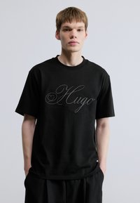 Μαύρο βαμβακερό μπλουζάκι με κοντά μανίκια, με μεγάλο καλλιγραφικό λογότυπο "Hugo" σε ασημί στο μπροστινό μέρος και στρογγυλή λαιμόκοψη.