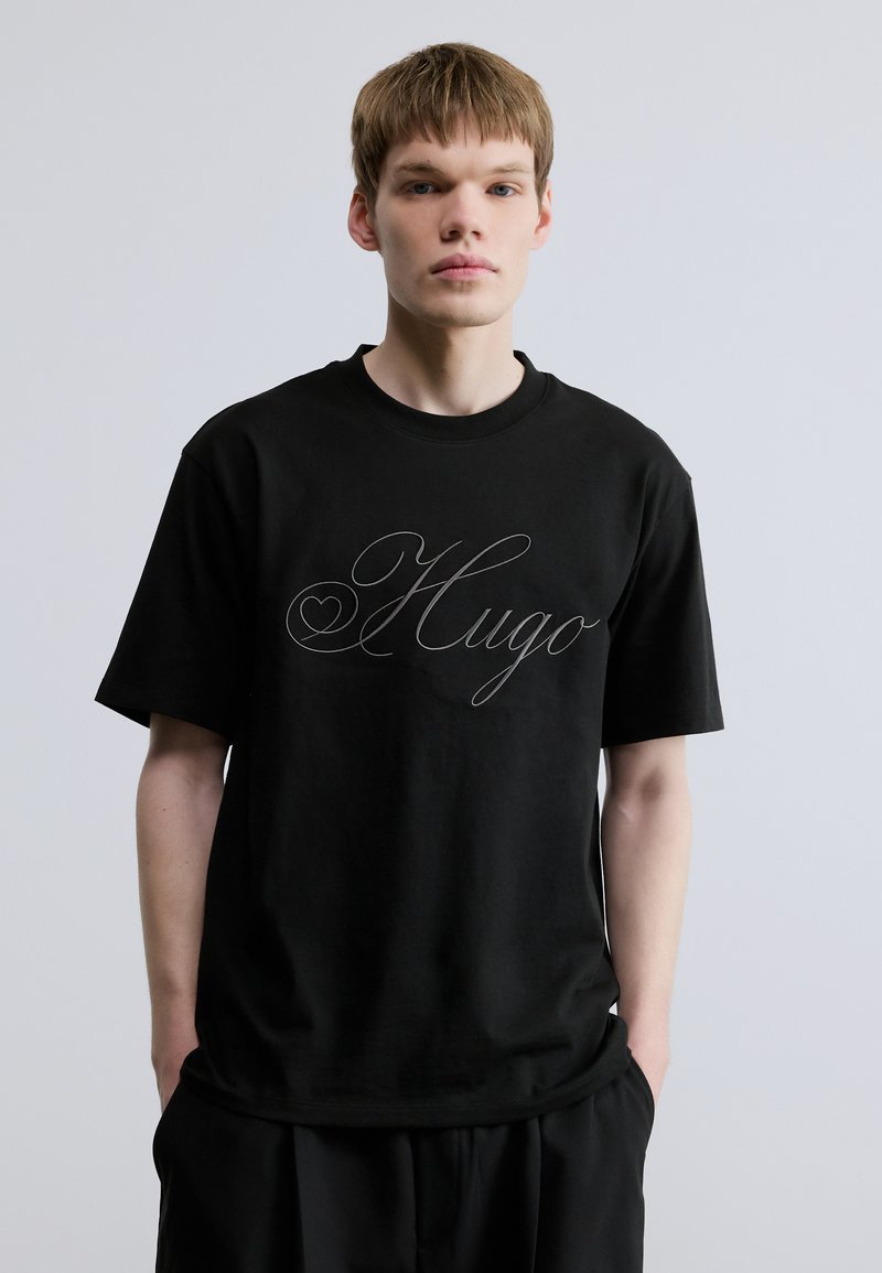 Μαύρο βαμβακερό μπλουζάκι με κοντά μανίκια, με μεγάλο καλλιγραφικό λογότυπο "Hugo" σε ασημί στο μπροστινό μέρος και στρογγυλή λαιμόκοψη.