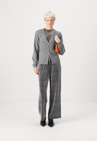 Hallja hall, millel on stringiga esiosa, kantud tekstuuriga musta pluusi peal, koos laia säärega hallide pükste ja mustade pahkluu saabastega.