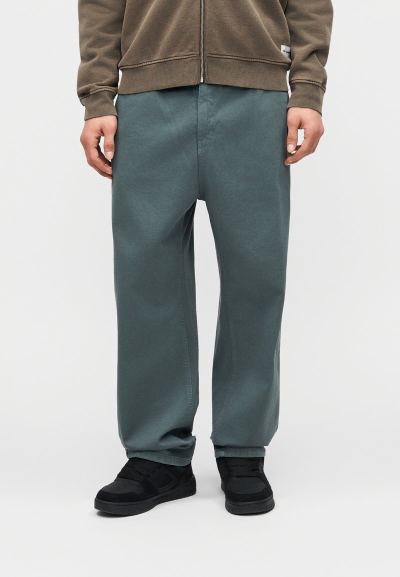 Person wearing muted green loose-fit pants, black sneakers, and a brown zip-up sweatshirt standing against a plain light background.