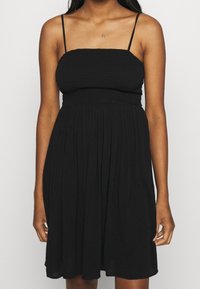 Černé bezramínkové šaty se smockovaným živůtkem, splývavou sukní a tenkými nastavovacími ramínky. Měkká, lehká tkanina s nařasenou strukturou.