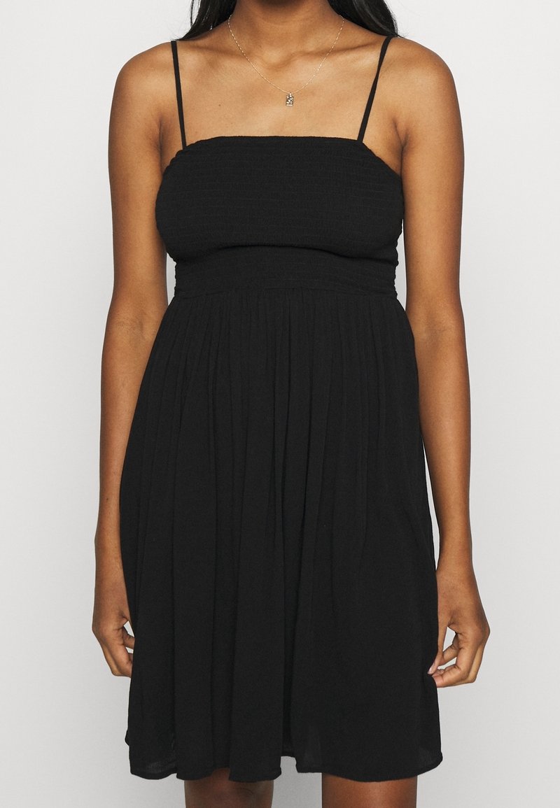 Černé bezramínkové šaty se smockovaným živůtkem, splývavou sukní a tenkými nastavovacími ramínky. Měkká, lehká tkanina s nařasenou strukturou.