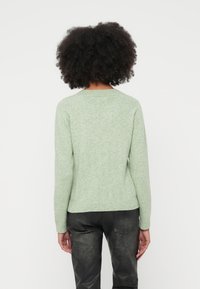 Vaaleanvihreä neulepaita, jossa on pyöreä pääntie ja pitkät hihat, pehmeä pinta ja istuva helma, yhdistettynä mustiin housuihin.