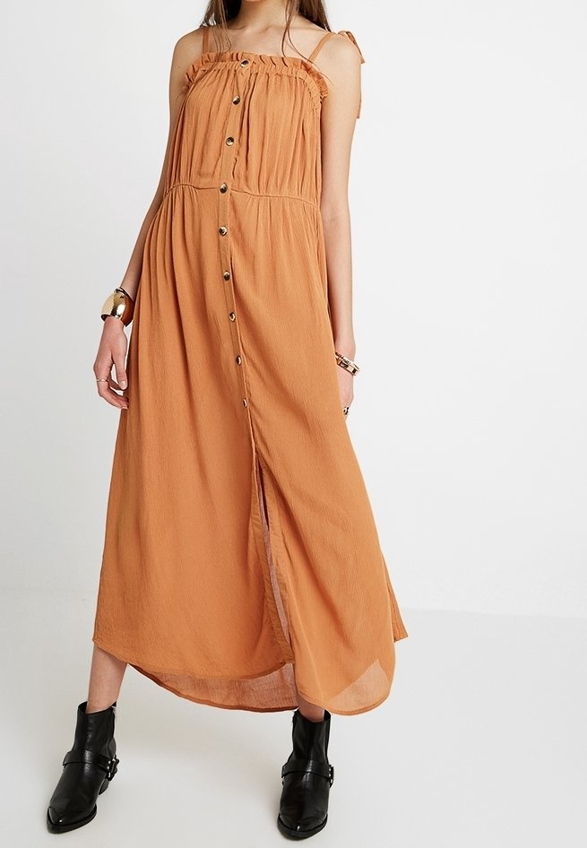 Kvinna klädd i en ärmlös, knappradad, rostorange midiklänning med volang på halsen och svarta ankelskor, står framför en vit bakgrund.