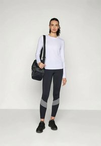 Kerge lilla pika varrukaga topp, millele on paigutatud tumedad retuusid hallide detailidega. Modell hoiab musta kotti ja kannab musti spordijalatsi.