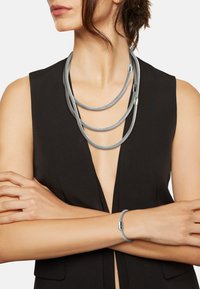 Grå flertrådet halskæde lavet af tekstureret materiale med metalknopper. Et matchende gråt armbånd har en elegant lås. Båret over en sort top.