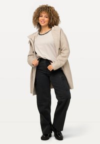 Abrigo de mezcla de lana gris claro con capucha, detalles de botones, camiseta gris y pantalones cargo negros. Modelo de pie con las manos en las caderas.