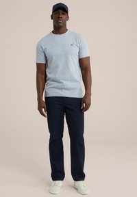 Tricou scurt de culoare gri deschis, cu un logo mic pe piept, asortat cu pantaloni bleumarin și adidași albi. Modelul stă în picioare.