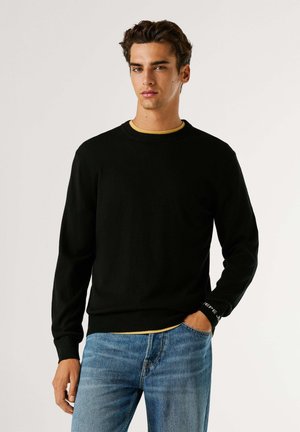 Maglione nero a maglia con scollo rotondo e maniche lunghe, caratterizzato da un orlo aderente. Indossato sopra una camicia gialla e abbinato a jeans blu.