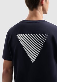 Man in een zwart T-shirt met een groot wit driehoekig ontwerp van diagonale lijnen op de achterkant, met zijn rug naar de camera gekeerd.