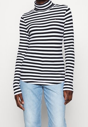 Personne portant un pull à col roulé rayé horizontal noir et blanc et un jean bleu clair, debout devant un fond uni.