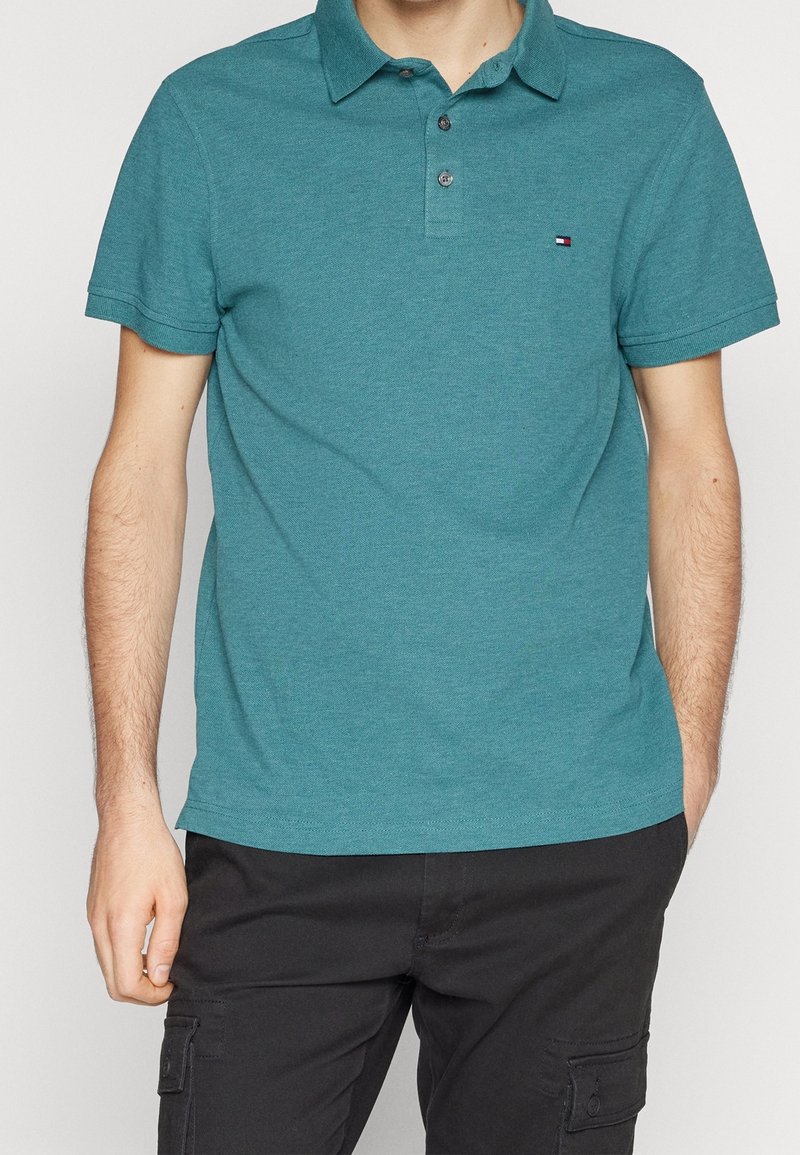 Homme portant un polo bleu sarcelle et un pantalon cargo noir, debout avec une main dans la poche sur un fond uni.