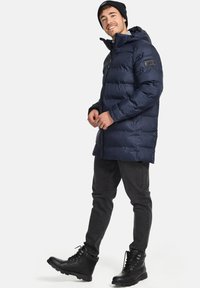 Chaqueta acolchada azul marino con capucha, cierre de cremallera y bolsillos laterales. Presenta un diseño acolchado y un parche con el logo en la manga. Combinada con jeans negros y botas.