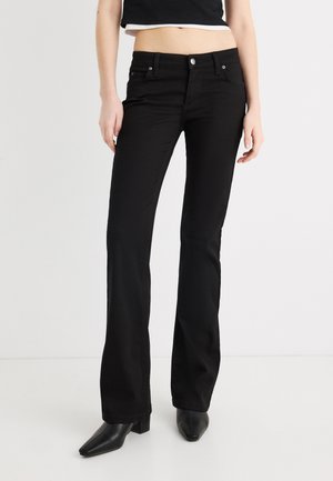 Kvinde iført sorte højtsiddende bootcut-jeans og sorte spidse støvler med hæl, stående foran en ensfarvet hvid baggrund.