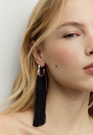 Plavuša svijetle puti koja nosi veliki srebrni obručasti naušnicu s dugim crnim resicama na jednostavnoj svijetloj pozadini.