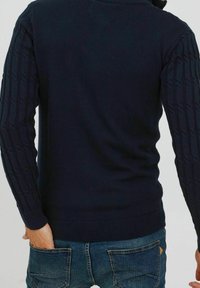Mann trägt einen dunkelblauen, strukturierten Pullover und Jeans, von hinten gezeigt, mit der rechten Hand in der Tasche vor einem schlichten Hintergrund.