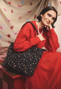 Moteris raudoname austame megztinyje ir tiulio sijone sėdi raudonoje suole, laiko juodą rankinę su žvaigždžių raštais ir perlų vėriniu prieš žvaigždėtą foną.