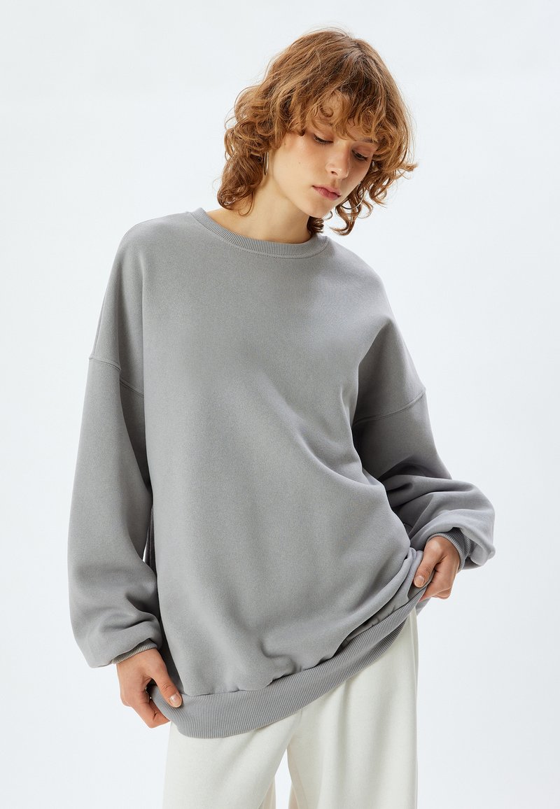 Felpa grigia con una vestibilità comoda, collo e polsini a coste e una texture morbida. Abbinata a pantaloni chiari e dal taglio rilassato.