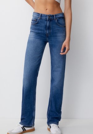 Person iført højtaljede blå jeans med lige ben og hvide sneakers med sorte striber, stående mod en ensfarvet hvid baggrund.