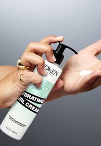 Vit plastflaska med en svart pump som är märkt "Fuktgivande Lockkräm." Kräm som dispenseras på en handflata, bakgrunden är grå.
