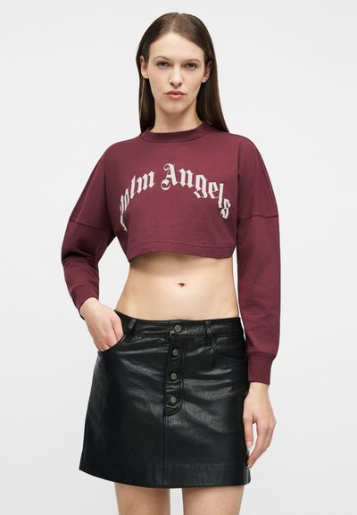 Γυναίκα που φοράει μπορντό cropped φούτερ με το κείμενο «Palm Angels» και μαύρη δερμάτινη μίνι φούστα με κουμπιά, πόζαρε με το ένα χέρι στην τσέπη.
