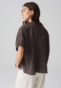 Camicia a maniche corte a righe marroni con vestibilità rilassata. Presenta un pannello posteriore solido a contrasto e spacchi laterali per un maggiore comfort.