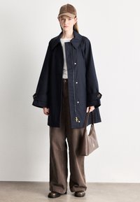 Ελαφρύ μπουφάν σε ναυτικό μπλε με φαρδιά γιακά, χρυσά κουμπιά με κλιπ και μανίκια σε σχήμα καμπάνας. Συνδυασμένο με καφέ παντελόνες φαρδιάς γραμμής και καφέ τσάντα.