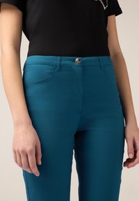 I pantaloni in misto cotone color teal presentano un design aderente, quattro tasche e una chiusura con bottone metallico, mostrati con una camicia nera.