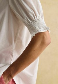 Arm, am Ellbogen gebeugt, trägt eine weiße Bluse mit elastischem, gerüschtem Bündchen und rosa Hosen, die am unteren Rand vor einem hellbeigen Hintergrund sichtbar sind.