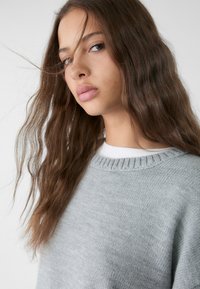 Helles graues Strickpullover mit einem gerippten Kragen, der eine weiche Textur und eine lässige Passform aufweist, getragen über einem weißen Hemd. Langes, welliges Haar umrahmt das Gesicht.