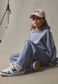 Sweat à capuche bleu clair et pantalon de survêtement assorti en tissu doux, avec des baskets blanches à accents violets et une casquette beige avec un logo.