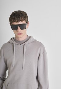 Sudadera gris claro con cordón, con una tela texturizada. El modelo lleva gafas de sol negras de gran tamaño, creando un fuerte contraste.