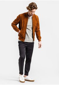 Braune Wildleder-Bomberjacke mit gerippten Bündchen, hellgrauer Pullover, dunkle Hose und weiße Sneakers mit sandfarbenen Sohlen. Das Modell steht.