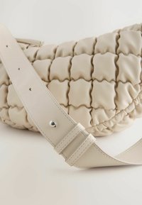 Beige håndtaske med en quiltet tekstur og blød, rund form. Har en bred læderrem med sølvhardware og justerbare huller.
