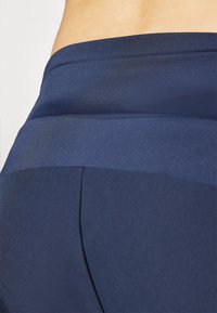Tamsiai mėlynos sportinės leggingės su lygiu paviršiumi, su besiūle juosmeniu ir kontrastingomis matinėmis bei blizgiomis audinio juostomis.