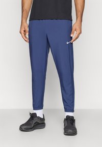 Άνδρας που φοράει μπλε σορτς Nike, μαύρα αθλητικά παπούτσια, λευκές κάλτσες και μαύρη μπλούζα, στέκεται μπροστά σε απλό φόντο.