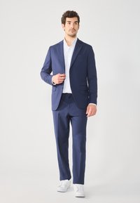 Traje azul marino de corte ajustado, con chaqueta de dos botones y pantalones a juego, acompañado de una camisa blanca y zapatillas blancas.