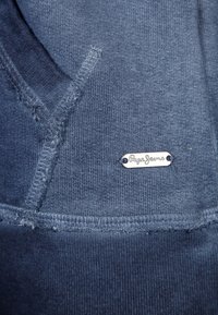 Blå bomullshoodie med synliga sömmar, ribbat fåll och en silveretikett med "Pepe Jeans"-logotypen. Mjuk textur och avslappnad design.