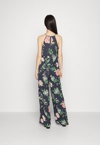 Macacão floral em azul-marinho com decote halter, apresentando padrões de flores em rosa e verde, e calças de perna larga. Costas abertas com um detalhe em formato de keyhole.