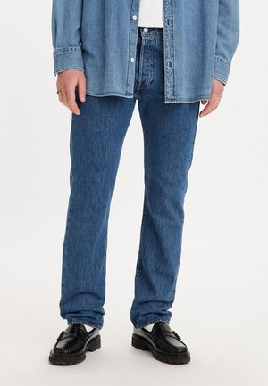 Jean en denim bleu avec une coupe droite, associé à une chemise à boutons bleu clair et des mocassins noirs avec des détails texturés.