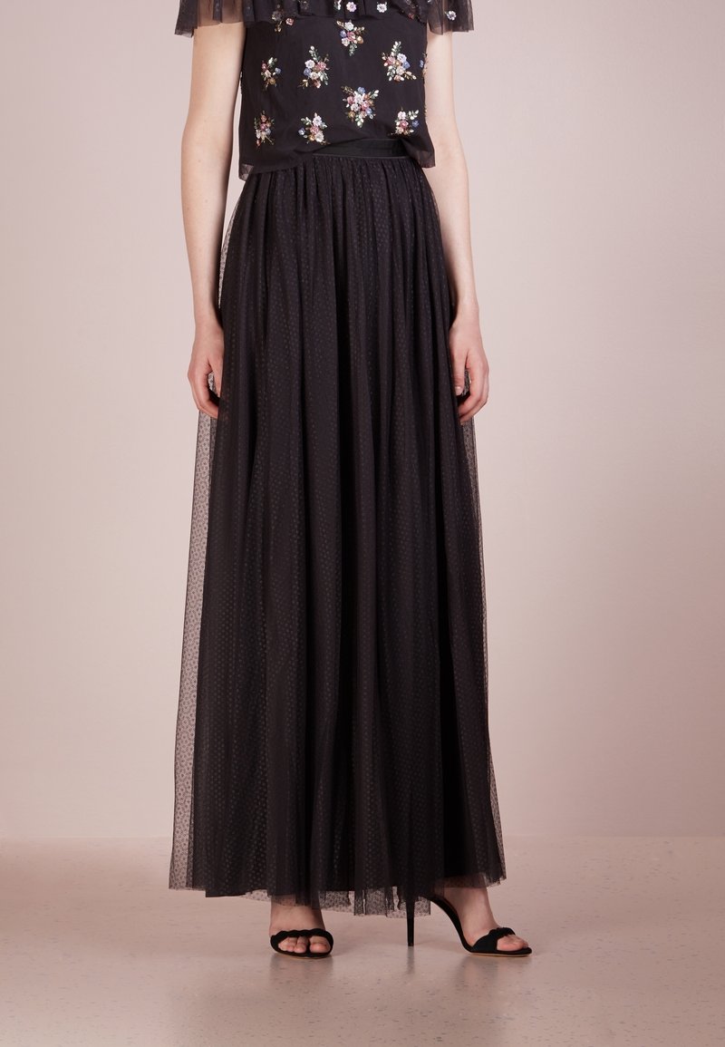 Jupe longue en tulle noir avec un motif à pois, présentant une taille froncée et un design superposé, associée à un haut brodé de fleurs et des talons noirs.