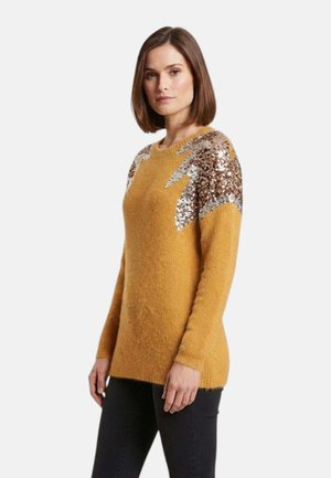 Donna con capelli castani lisci che indossa un maglione giallo senape con dettagli di fulmini in paillettes argento e bronzo sulle spalle.