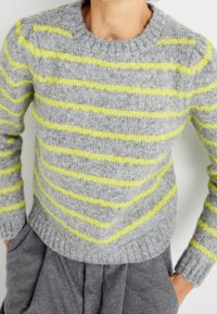 Harmaa villapaita keltaisilla raidoilla, pyöreä kaulus, resorit hihansuissa ja helmassa. Pehmeä rakenne ja istuva malli, puettu harmaiden housujen kanssa.