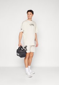 Beige oversized t-shirt with graphic prints, paired with beige shorts. Holding a black Adidas duffel bag. Light athletic shoes and white socks.