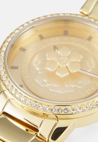 Orologio d'oro con quadrante rotondo, dial d'oro testurizzato, accenti cristallini attorno al bracciale e lancette delle ore e dei minuti minimaliste.