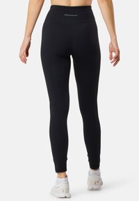 Leggings neri a vita alta realizzati in tessuto elasticizzato, con una superficie liscia e un logo sulla cintura. Indossati con scarpe sportive bianche.