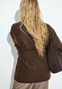 Brauner Wollmischung-Wickelmantel mit einem Gürtel, der eine weiche Textur und eine taillierte Passform aufweist. Eine dunkle Lederhandtasche hängt über der Schulter.