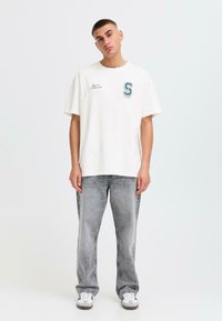 White cotton t-shirt with a round neck, short sleeves, featuring a green embroidered letter 'S' on the left chest. Paired with faded gray jeans.