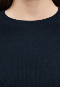 Λαιμός και άνω μέρος στήθους ενός ατόμου που φοράει σκούρο μπλε πλεκτό πουλόβερ με στρογγυλή λαιμόκοψη, με μακριά καστανά μαλλιά εν μέρει ορατά.